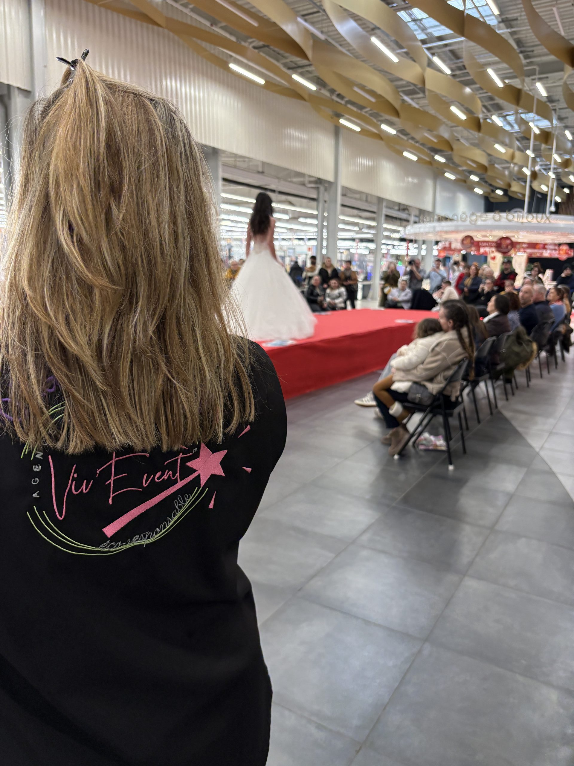 Membre de l’équipe Viv’Event de dos devant un défilé en robe de mariée, dans la galerie d’un centre commercial